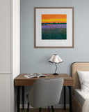 Framed artwork above a desk with a chair and lamp in a room setting.