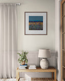 Framed artwork above a wooden bench with a lamp and plant in a room with curtains and a door.