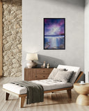 A framed vertical landscape art print featuring a purple and blue sky over a reflective body of water, displayed on a light gray wall above a wooden daybed in a modern, rustic living room setting.