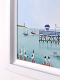 A framed art print depicting a coastal harbor scene with seagulls on rocks in the foreground, small boats on the water, a pier, and a lighthouse in the distance, shown against a white wall.
