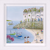 A framed coastal village painting featuring white houses on a grassy cliff overlooking a blue bay, with small boats, a sandy beach, and people engaging in beach activities. The artwork is presented in a white square frame.