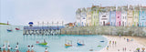 A colorful coastal village scene painting with pastel buildings along the shore, a pier extending into calm blue water, small boats, and beachgoers on the sand.