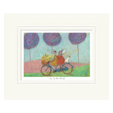 Framed artwork of a couple on a bicycle with a dog, under purple trees, titled 'Joy to the World'.