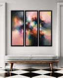 A three-panel framed abstract wall art set hangs above a light gray bench with wooden legs in a styled interior. The artwork features soft blended tones of pink, blue, orange, yellow, and dark charcoal, with no visible brand text.
