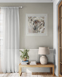 A styled interior corner with a light gray wall, white curtains, a framed abstract artwork, a wooden bench, a potted plant, books, and a textured table lamp.