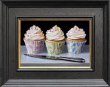 Three cupcakes with white frosting and colorful wrappers on a reflective surface, framed in black.