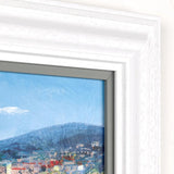 A close-up of a framed art print showing an abstract cityscape with a mountain under a blue sky, set in a white textured frame.