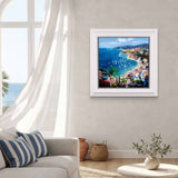 A framed coastal landscape painting hangs on a light gray wall in a bright living room. The artwork depicts a sunny seaside town with blue ocean water, boats, palm trees, and colorful buildings.