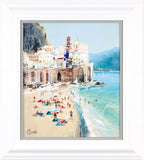A framed coastal art print depicting a sunny beach scene with colorful buildings, swimmers, and beachgoers, presented in a white frame with a light gray mat.