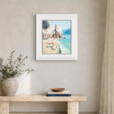 A framed art print depicting a colourful beach town scene in Italy, hanging on a beige wall above a stone console table with a vase and books.