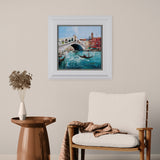 framed cityscape artwork of venice water with boat and buildings surrounding on plain cream wall above a wooden chair and metal side table