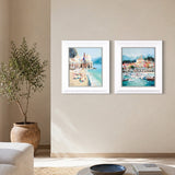 Two framed coastal art prints hanging side by side on a beige wall in a styled living room setting with a potted olive tree and a sofa.