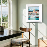 A framed art print depicting a colorful coastal harbor town with boats and mountains, displayed on a light sage green wall in a modern room setting with a wooden table and chair.