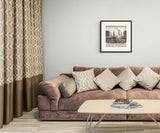 A styled living room scene featuring a mauve-brown velvet sofa with beige and gray patterned throw pillows, a light wood coffee table, patterned curtains, and a framed art print on a light gray wall.