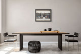 A modern dining room setup featuring a rectangular wooden table with a black metal frame, two black chairs with chrome legs, a black woven pouf, and a framed art print on a light gray wall.