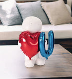 White figurine holding a red heart-shaped balloon and a blue 'U' balloon on a wooden surface with a couch in the background.