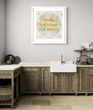 A framed vintage-style world map print with the text 'FORTUNE FAVOURS THE BRAVE' in yellow, displayed on a light gray wall above a rustic kitchen counter with wooden cabinets and a white farmhouse sink.