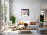 A colorful abstract framed wall art print hangs on a white wall in a bright, modern living room with a white sofa, yellow and pink pillows, a wooden coffee table, and potted plants.