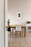 A framed art print featuring a dark blue and black architectural scene with a bridge, displayed on a light beige wall above a wooden dining table and chairs in a modern, minimalist room.