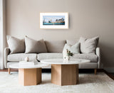 A framed art print depicting fishing boats on the water, displayed on a beige wall above a modern light gray sofa in a styled living room setting.