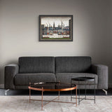 A framed art print depicting a cityscape with tall buildings and smokestacks, displayed on a gray wall above a modern dark gray fabric sofa with two round coffee tables in front.