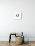 A small framed art print depicting a colorful boat scene, styled on a white wall above a wooden floor with a stool, a woven basket, and two glass bottles.