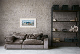 A framed art print depicting a blue and white helicopter on water, displayed on a textured gray brick wall above a brown velvet sofa in a modern living room setting.