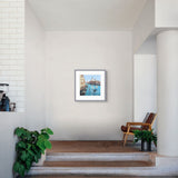 A framed art print depicting a scenic canal with blue water and warm-toned buildings, displayed on a light gray wall in a modern, minimalist interior setting with a wooden chair and potted plants.