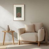 A framed art print of a small brown dog on a dark green background, styled on a beige wall above a cream armchair and a wooden side table.
